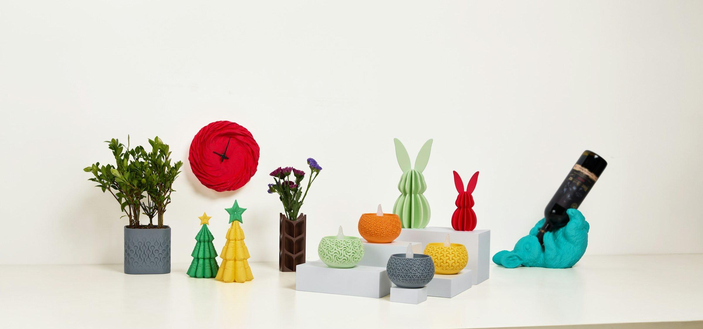 Peças decorativas impressas em 3D sobre uma bancada branca: vasos, árvores de Natal, coelhos, porta-velas, um relógio de parede e um porta-garrafas.